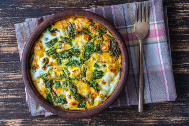 Ceramic bowl with vegetable frittata, simple vegetarian food. Frittata with egg, pepper, onion, cheese and green wild garlic leaves on table, close up, copy space. Healthy egg omelet