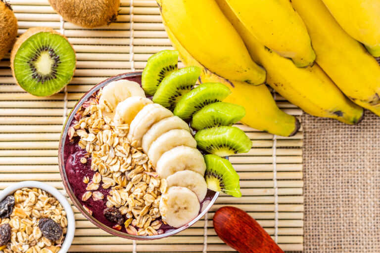 Acai bowl with kiwi, banana and oat flakes on a table with tropical decor in top view.